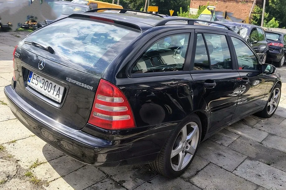 Czarny samochód marki Mercedes-Benz w nadwoziu kombi, zaparkowany na betonowym podjeździe. Widoczny jest tył i prawy bok pojazdu. Samochód posiada tablicę rejestracyjną o numerze ES 3001A oraz napis KOMPRESSOR na klapie bagażnika. Na dachu znajdują się czarne relingi. W tle widoczne są inne zaparkowane samochody oraz fragmenty zieleni i budynku.