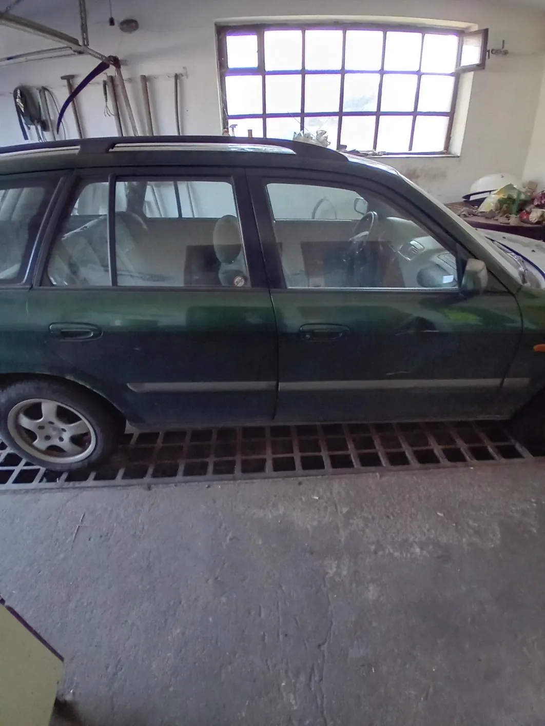 Zielony samochód typu kombi w garażu. Samochód stoi na metalowej kratce, w tle duże okno z kratą i narzędzia wiszące na ścianie. Podłoga garażu jest betonowa, po prawej stronie widoczne są różne przedmioty i biały kask.