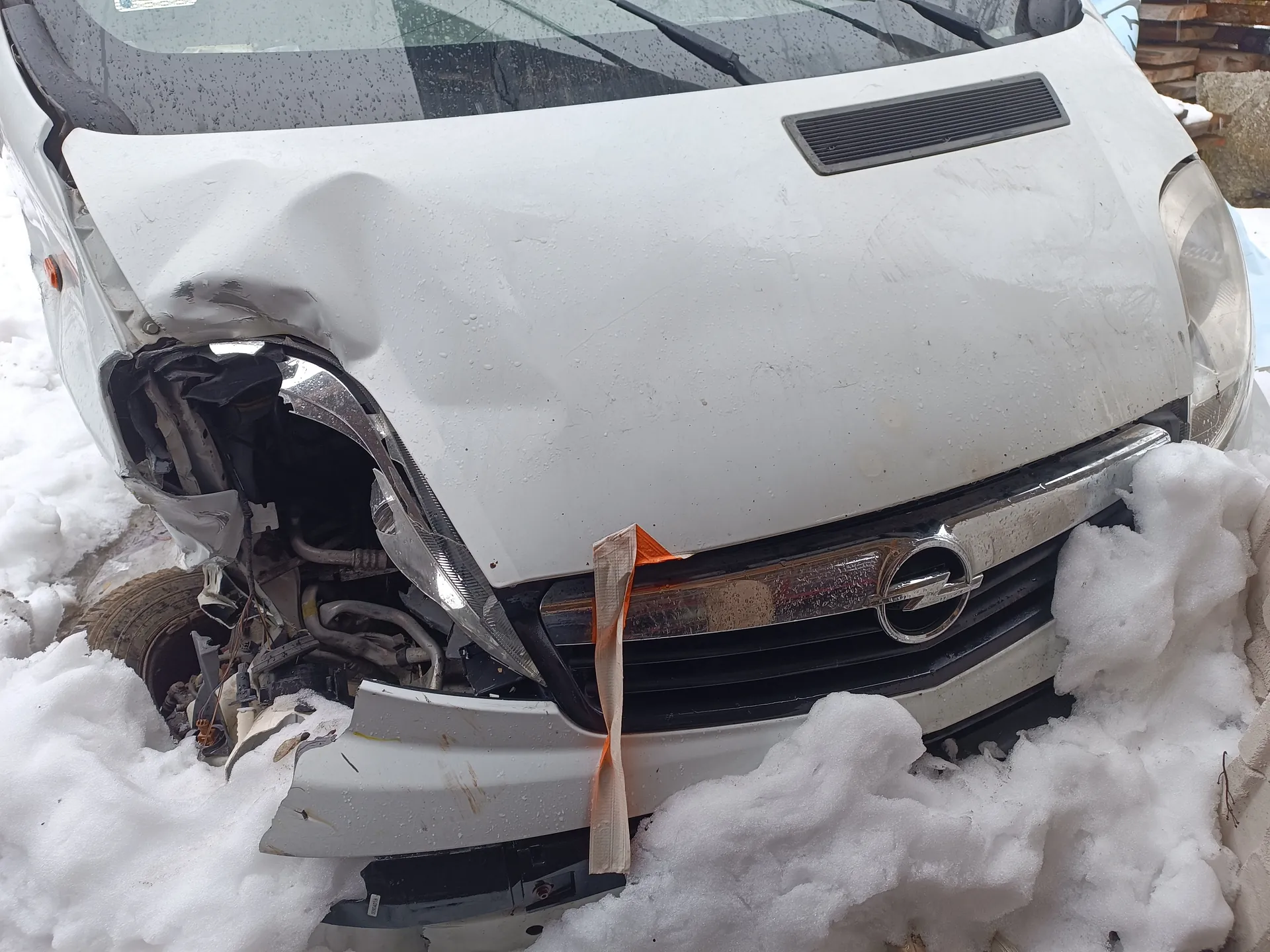 Biały samochód marki Opel z uszkodzonym przodem po kolizji. Widoczne zgniecione elementy nadwozia po lewej stronie, uszkodzony reflektor, odsłonięte części silnika oraz pomarańczowa taśma przytwierdzona do pasa przedniego. Samochód znajduje się na śniegu, w tle widoczne są stosy materiałów budowlanych. Na masce i szybie deszczu widoczne krople wody.