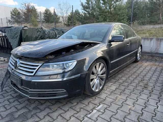 Ciemnoszary samochód osobowy marki Volkswagen z otwartą maską i uszkodzonym przodem, stojący na brukowanej powierzchni. W tle widoczne są drzewa, ogrodzenie oraz zadaszony obiekt z zielonymi pokrowcami. Niebo częściowo zachmurzone.