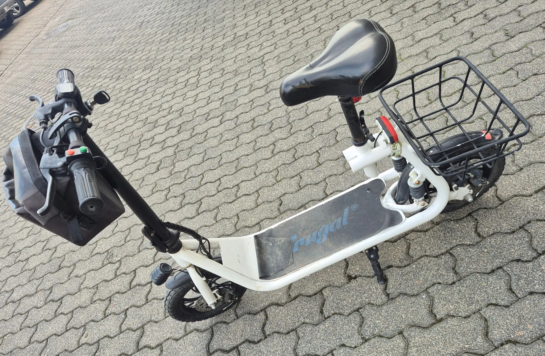 Biały skuter elektryczny z czarnym siedzeniem i czarnym koszem z przodu, zaparkowany na kostce brukowej. Na podest dla stóp widnieje niebieski napis 'ugal'. Pojazd posiada czarną kierownicę z zamontowaną torbą oraz dwa małe koła.