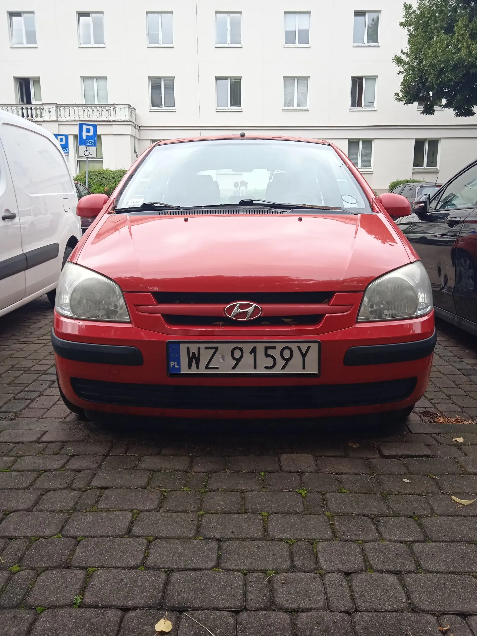 Czerwony samochód marki Hyundai zaparkowany przodem do widza na kostce brukowej. Pojazd posiada polską tablicę rejestracyjną o numerze WZ 9159Y. W tle widoczny jest biały, wielopiętrowy budynek z licznymi oknami oraz fragmenty innych samochodów: białego vana po lewej i czarnego auta po prawej.