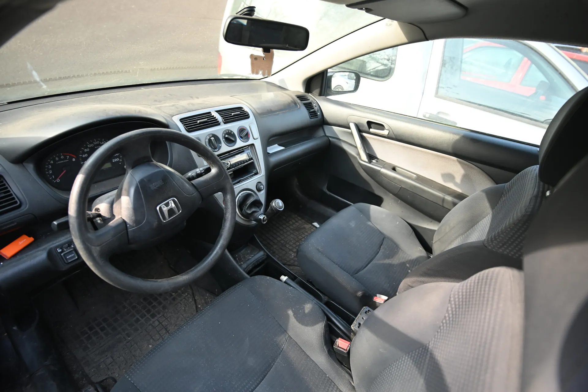 Wnętrze samochodu marki Honda z widocznym kierownicą z logo Honda, tablicą rozdzielczą z licznikami prędkości i obrotomierzem, centralną konsolą z odbiornikiem radiowym i klimatyzacją, czarnymi siedzeniami oraz lusterkiem wstecznym. Na podłodze widoczne są czarne dywaniki. Przez przednią szybę widać fragment innego pojazdu. Wnętrze wydaje się nieco zapylone.