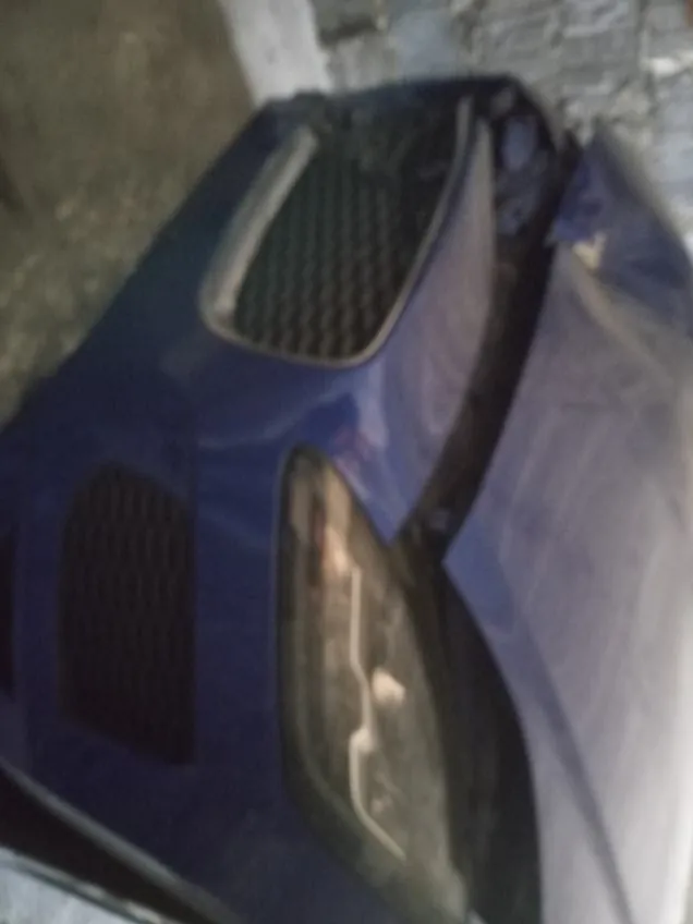 Niebieski samochód z otwartą maską, widok przedniej części z atrapą chłodnicy w kształcie siatki, reflektorem i tłem z kamienną ścianą oraz betonową powierzchnią.