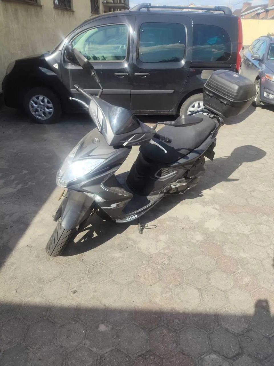 Czarny skuter marki KYMCO zaparkowany na chodniku z sześciokątnymi płytkami. Za skutrem widoczny ciemnoszary samochód typu van oraz częściowo widoczny inny pojazd. W tle budynek z beżową ścianą i oknami. Scena na otwartym powietrzu w świetle dziennym.