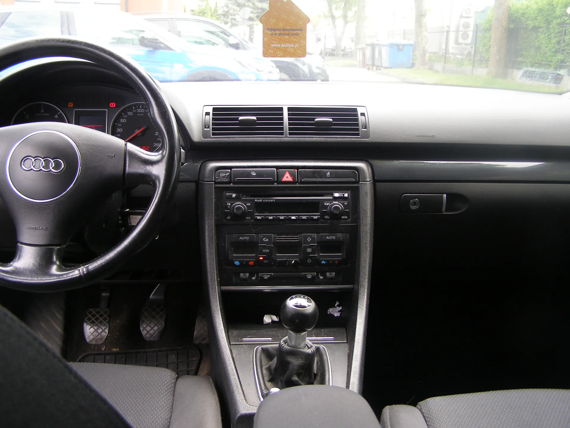 Wnętrze samochodu marki Audi z widocznym kierownicą z logo Audi, wskaźnikami prędkości i obrotomierzem na desce rozdzielczej. W centrum konsoli środkowej znajduje się radio z przyciskami, w tym czerwonym trójkątem ostrzegawczym, oraz dźwignia zmiany biegów. Na lusterku wstecznym wiszący jest żółty znaczek z napisem: „Najlepsze ubezpieczenie w atrakcyjnej cenie! www.ubezpieczenia.pl”. Przez przednią szybę widoczne są zaparkowane samochody, drzewa i niebieski pojemnik na śmieci. Siedzenia i deska rozdzielcza są w kolorze czarnym.