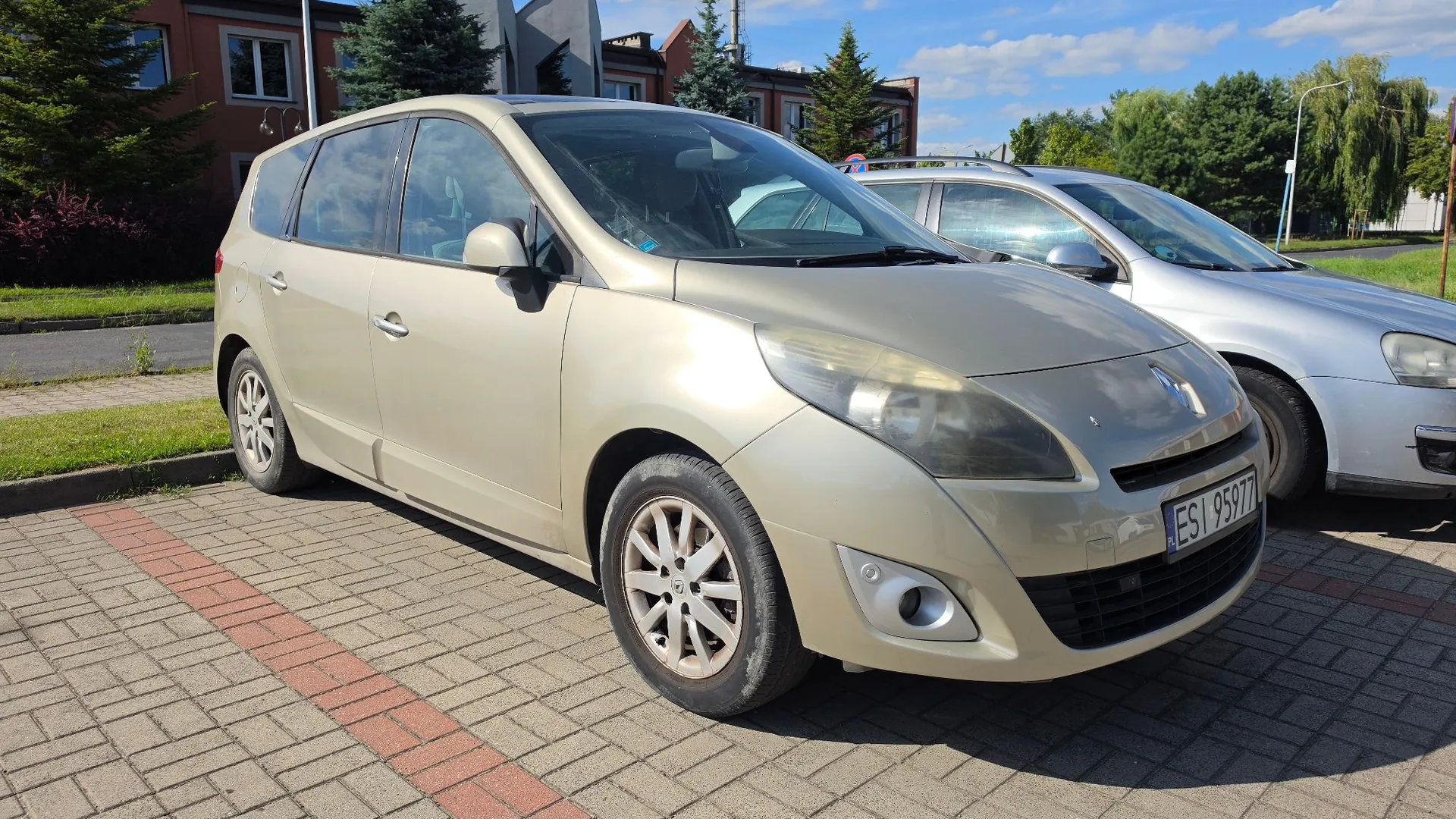 Jasnozłoty samochód osobowy marki Renault Scenic zaparkowany na kostce brukowej. Pojazd jest ustawiony pod kątem, widoczny z przodu i z lewej strony. Na samochodzie znajduje się tablica rejestracyjna o numerze ESI 95977. W tle widoczny jest inny, srebrny samochód, zielone krzewy, drzewa oraz fragmenty budynków mieszkalnych pod niebieskim niebem z lekkimi chmurami.