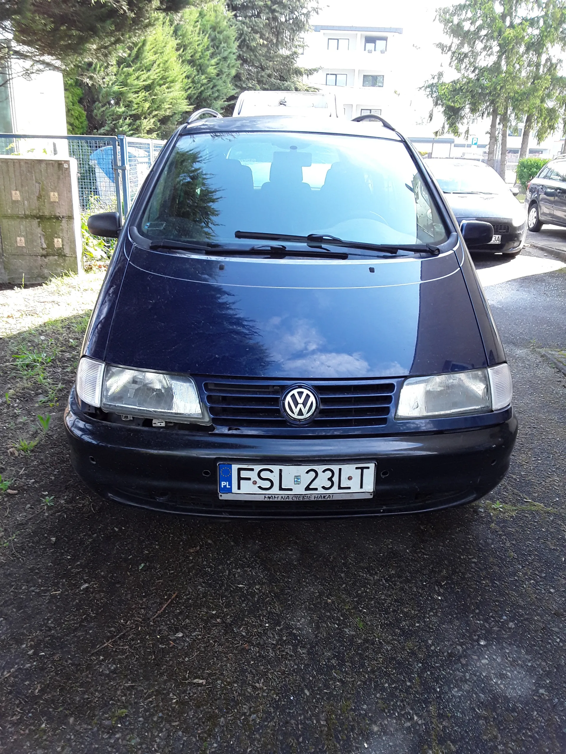 Ciemnoniebieski samochód marki Volkswagen zaparkowany na podłożu z żwiru. Widoczna tablica rejestracyjna z polskim kodem 'PL' i numerem 'FSL 23LT', poniżej napis 'MAM NA CIEBIE OKO'. W tle inne zaparkowane pojazdy, drzewa, budynek oraz ogrodzenie.