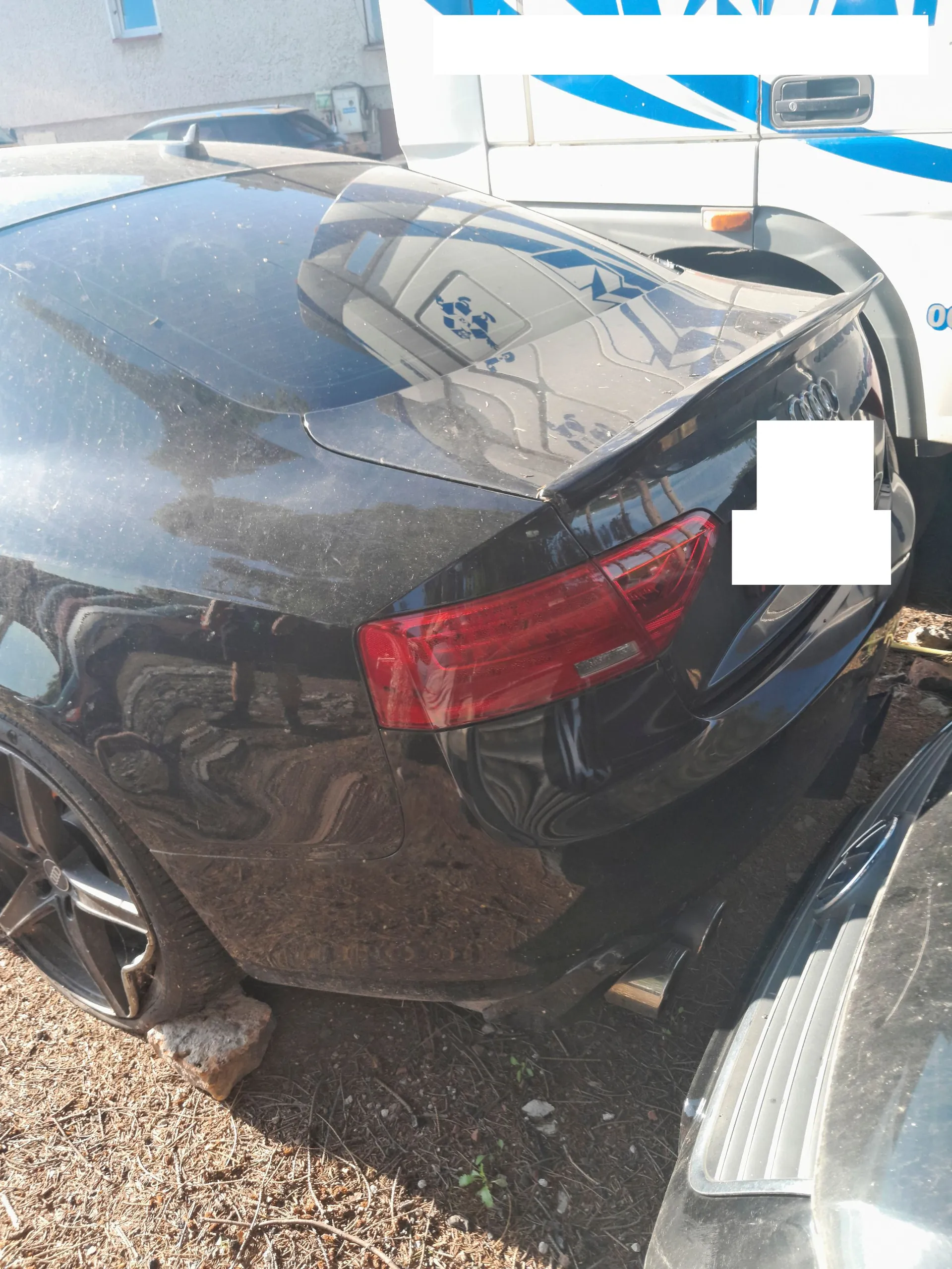 Tylna część czarnego samochodu marki Audi, widoczna z perspektywy trzech czwartych. Samochód jest zakurzony, posiada tylny spojler oraz czerwone światło stopu. Część tylnej klapy jest zakryta białym prostokątem. Pojazd stoi na piaszczystym podłożu obok innego ciemnego samochodu i białego vana.