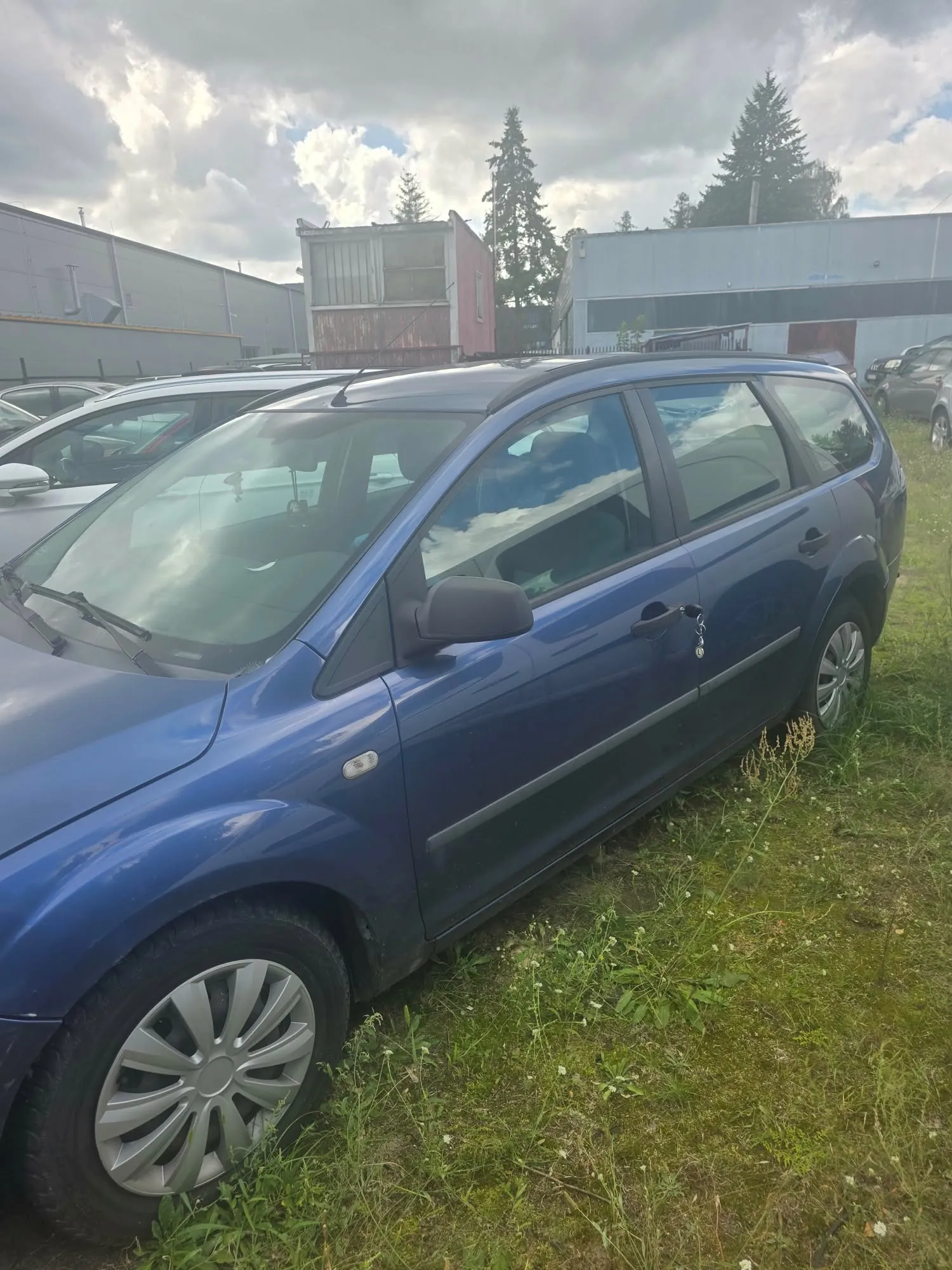 Niebieski samochód typu kombi zaparkowany na trawiastym poboczu. W tle widoczne są inne zaparkowane pojazdy, przemysłowe budynki oraz drzewa iglaste pod zachmurzonym niebem.