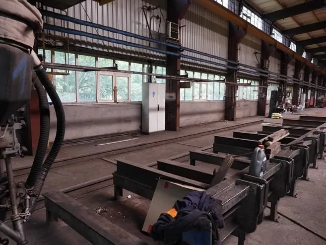 Wnętrze hali przemysłowej lub warsztatu. W pomieszczeniu znajdują się metalowe konstrukcje i ramy ułożone w rzędach na posadzce. W tle widoczna jest długa ściana z rzędem okien, przez które wpada światło dzienne. Wzdłuż sufitu i ścian biegną niebieskie rury oraz stalowe belki konstrukcyjne. Po prawej stronie stoi biała szafka metalowa, a na jednej z ram leży szara butelka z płynem oraz ciemne ubranie.