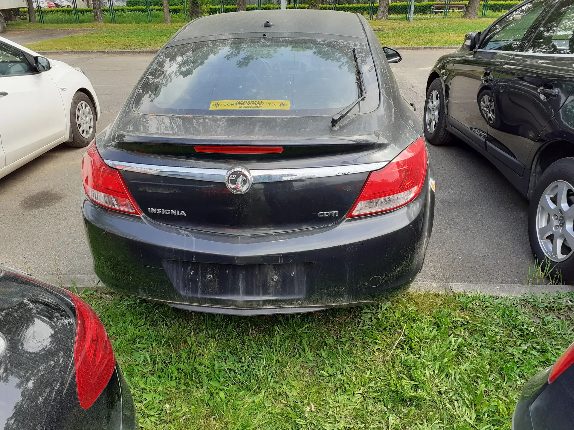 Tylny widok czarnego samochodu marki Opel Insignia zaparkowanego na trawiastym poboczu między białym a czarnym samochodem. Na tylnej szybie znajduje się żółta naklejka z napisem 'MARSHALL CONSTRUCTION LTD'. Na klapie bagażnika widoczne są napisy 'INSIGNIA' oraz 'CDTI' oraz logo marki Opel.