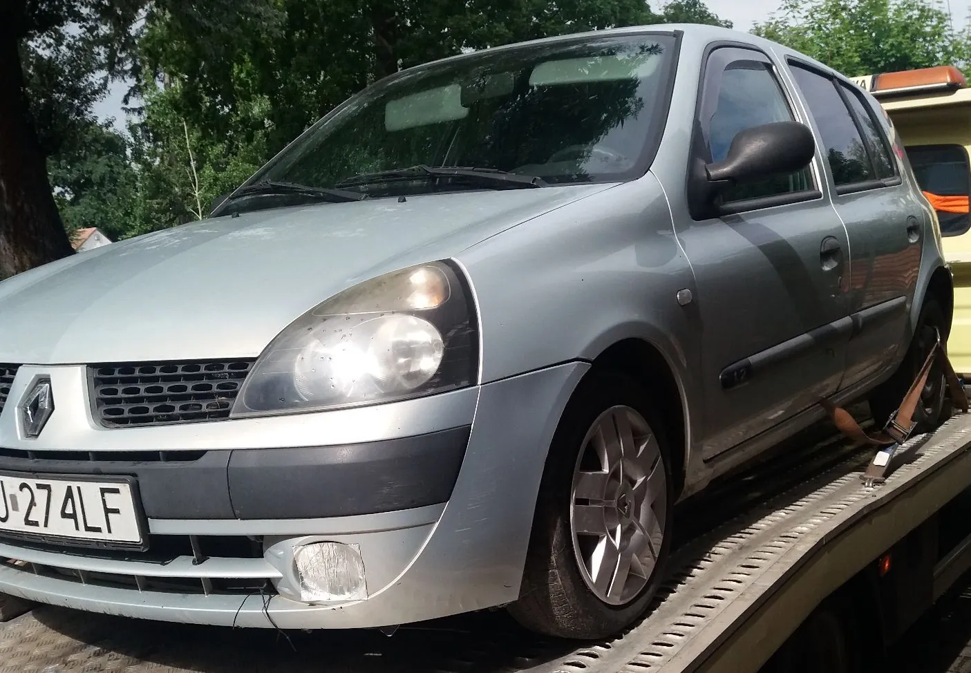 Srebrny samochód marki Renault Clio zaparkowany na lawecie. Widoczny jest przód i bok pojazdu. Samochód posiada tablicę rejestracyjną o numerze J 274LF. W tle znajdują się zielone drzewa.