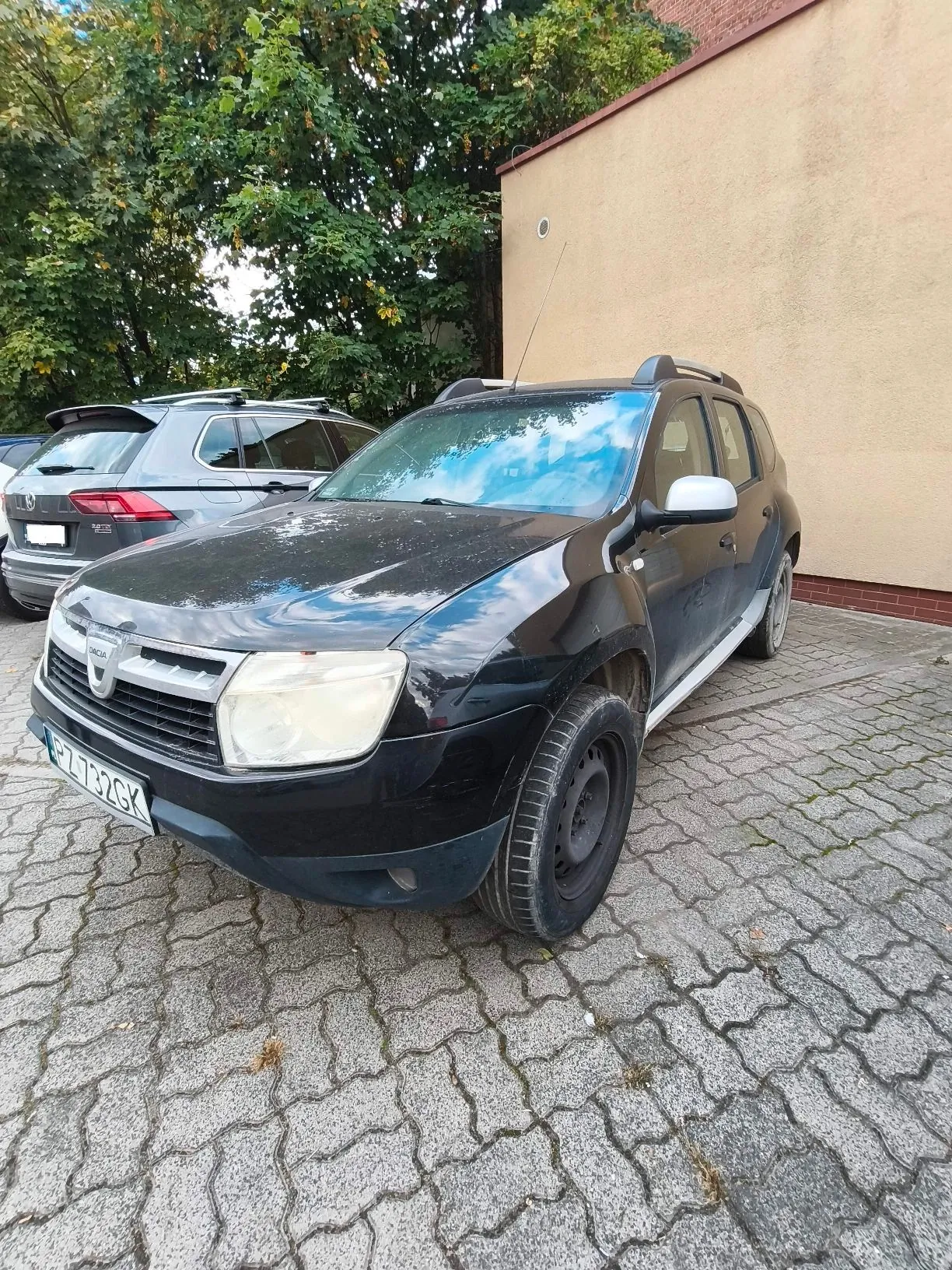 Czarny samochód marki Dacia zaparkowany na kostce brukowej przed jasną ścianą budynku. Obok stoi srebrny samochód. W tle widoczne są zielone drzewa. Samochód ma tablicę rejestracyjną o numerze PZ 732GK.