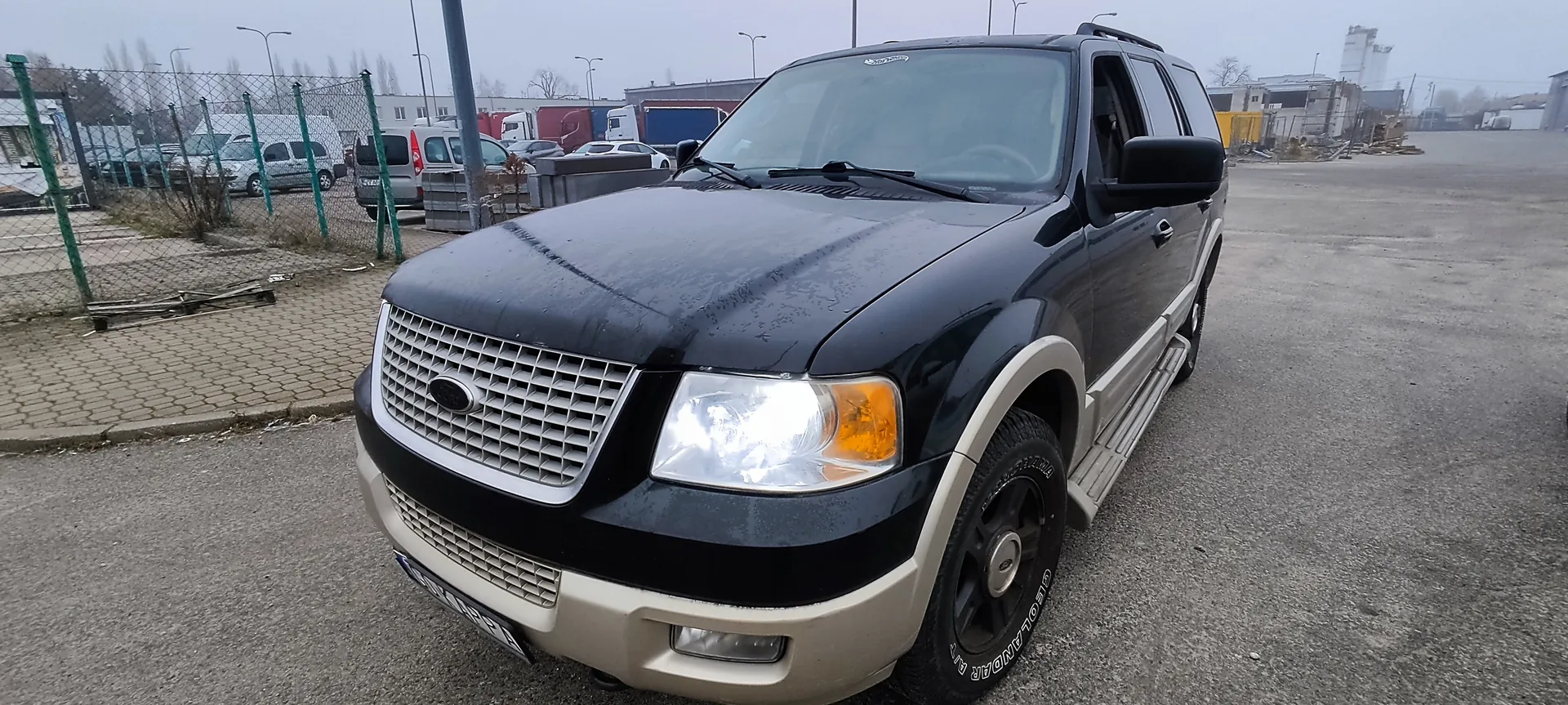 Czarny i beżowy SUV marki Ford zaparkowany na asfaltowym placu. W tle siatka ogrodzeniowa z zielonymi słupkami, inne pojazdy, budynki przemysłowe oraz pochmurne niebo. Widoczne są reflektory, atrapa chłodnicy z logo Ford oraz opony z napisem GEOLANDAR.