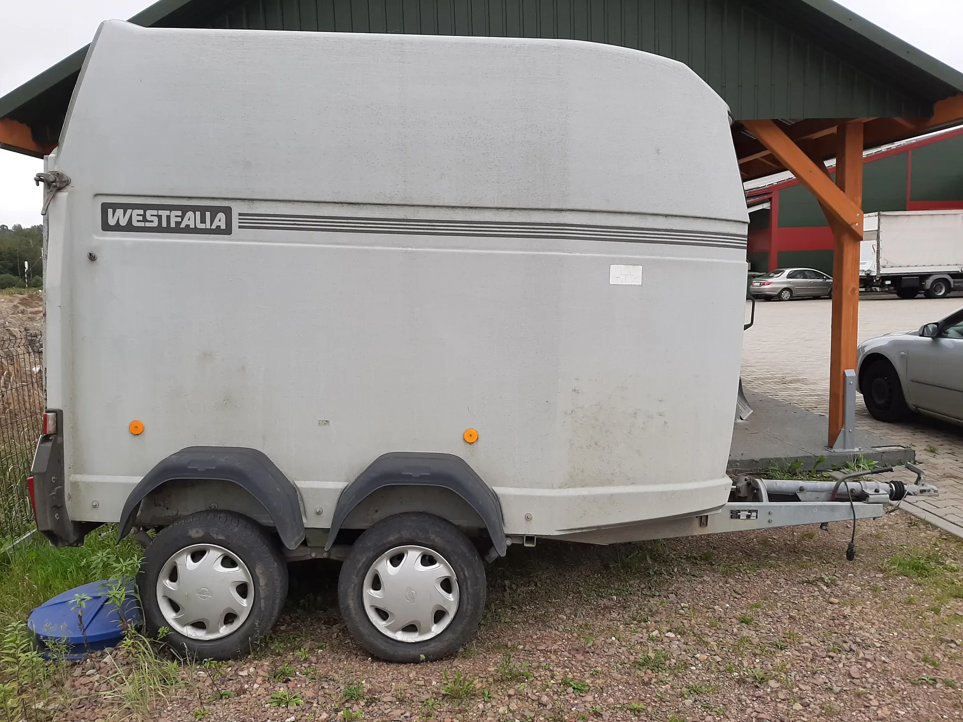 Szary przyczepa marki WESTFALIA z czarnymi osłonami kołami, srebrnymi felgami i pomarańczowymi reflektorami. Przyczepa stoi na żwirowym podłożu z trawą, w tle zielony budynek z drewnianymi słupami, samochody i ciężarówka. Na przyczepie widoczny czarny napis WESTFALIA oraz trzy poziome czarne linie.