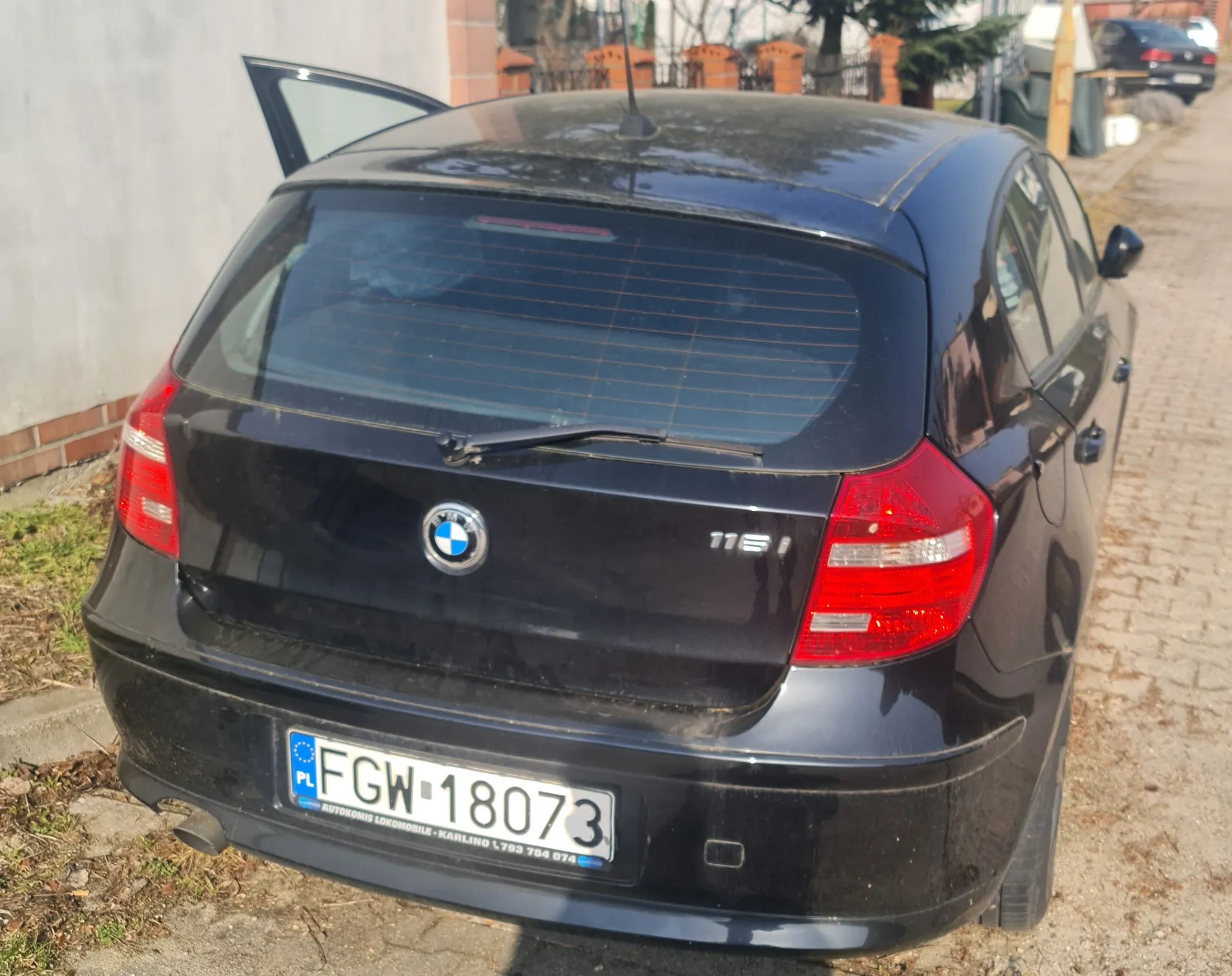 Czarny samochód marki BMW model 116i widziany od tyłu. Otwarte lewe tylne drzwi. Tablica rejestracyjna z białym tłem i czarnym tekstem: FGW 18073, po lewej stronie niebieski pasek z literą PL oraz napisem AUTOKOMIS LOKOMOBILE - KARLINO 1 793 754 074. Czerwone światła tylnego, logo BMW na bagażniku. Samochód zaparkowany na brukowanej drodze obok białej ściany z ceglanym podłożem. W tle widoczne są inne pojazdy, ogrodzenie i drzewa.