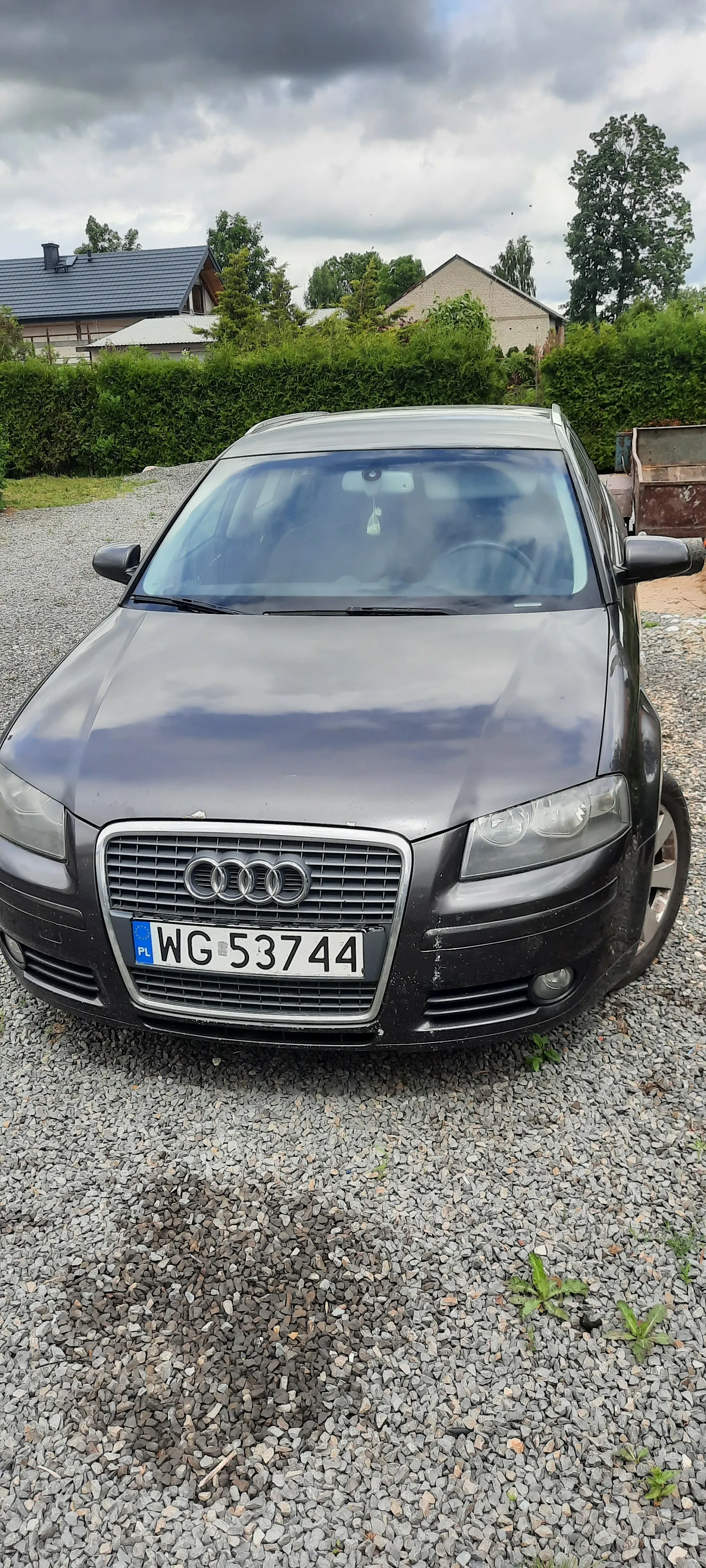 Czarny samochód marki Audi zaparkowany na żwirowym podjeździe. Widoczna tablica rejestracyjna z numerem WG 53744 oraz oznaczeniem PL. W tle dom z szarą dachówką, zielone krzewy, drzewa i chmurne niebo.