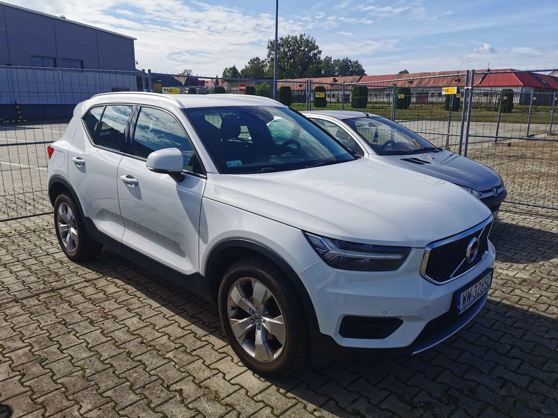 Biały samochód marki Volvo XC40 zaparkowany na kostce brukowej. Obok niego stoi srebrny samochód. W tle widoczny jest metalowy płot, budynki przemysłowe oraz zielone krzewy pod jasnym, lekko zachmurzonym niebem. Samochód ma tablicę rejestracyjną zaczynającą się od liter WW.