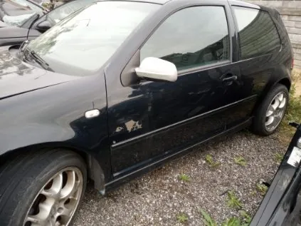 Czarny samochód osobowy typu hatchback zaparkowany na żwirowym podłożu. Widoczne uszkodzenia lakieru na boku pojazdu oraz biały lusterko boczne. W tle częściowo widoczny inny samochód i fragment ogrodzenia. Podłoże pokryte drobnym żwirem z nielicznymi zielonymi roślinami.