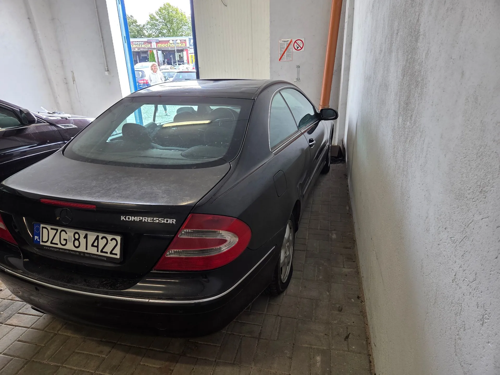 Czarny samochód osobowy marki Mercedes z napisem KOMPRESSOR na klapie bagażnika, zaparkowany w białym garażu lub warsztacie. Pojazd ma polską tablicę rejestracyjną o numerze DZG 81422. W tle widoczne jest otwarte wejście do budynku, przez które widać fragmenty ulicy i szyld z napisem 'mechanika'.