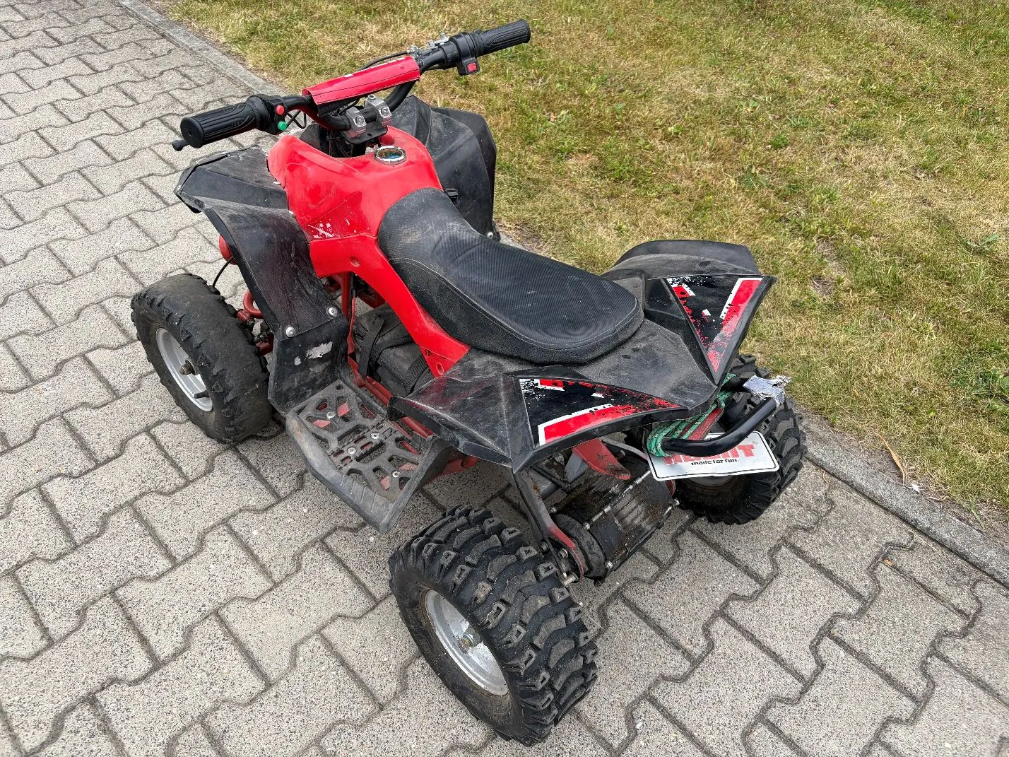 Czerwono-czarny quad dziecięcy zaparkowany na szarej kostce brukowej. Pojazd ma szerokie, terenowe opony, czarne błotniki i siedzenie. W tle widoczny jest pas zielonej trawy.