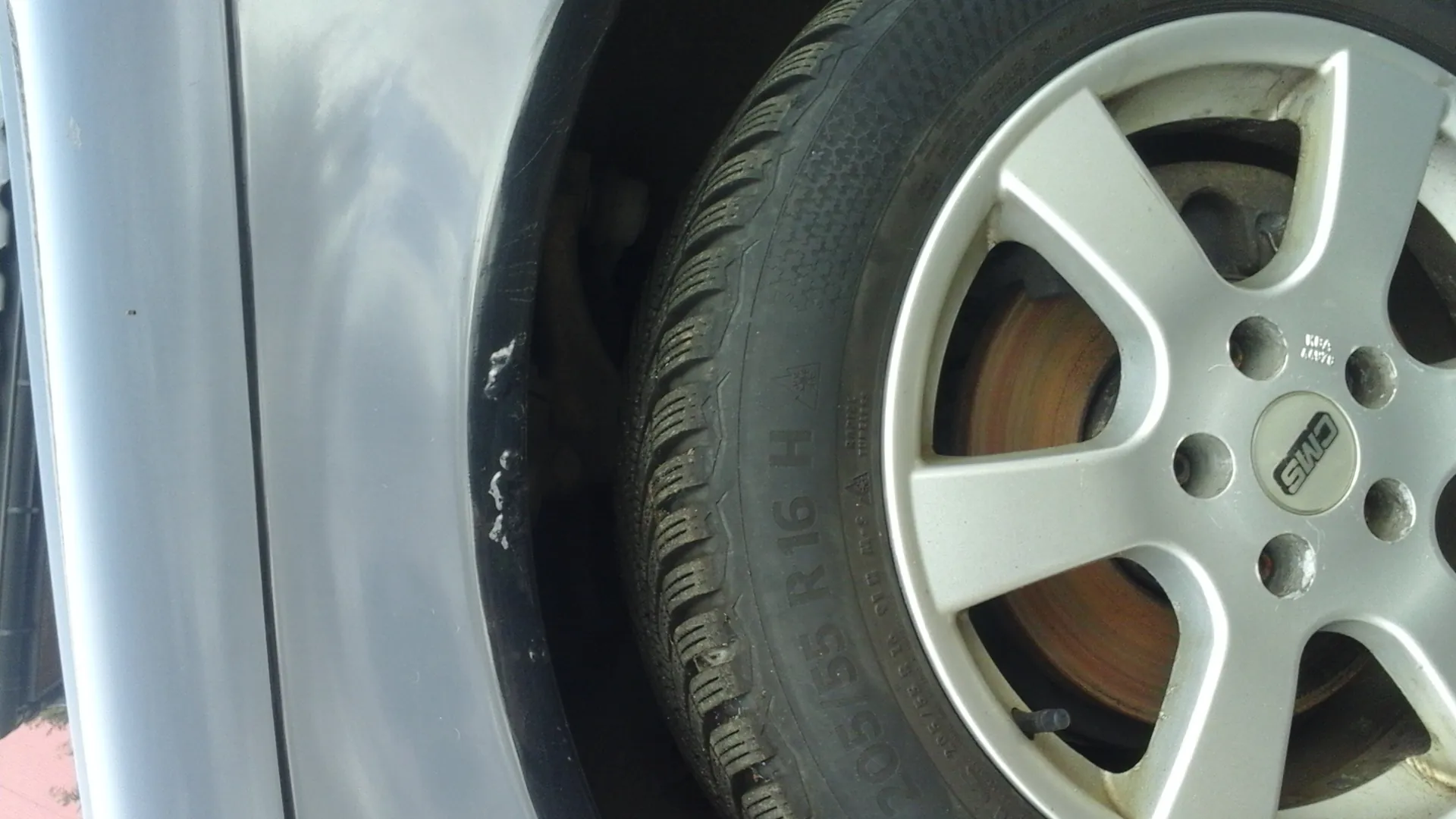 Zbliżenie na koło samochodu z srebrnym felgiem oznaczonym logo 'CMS'. Na oponie widoczny napis '205/55 R16 H' oraz inne dane techniczne. Widoczna część srebrnego nadwozia samochodu i tarcza hamulcowa za felgą.