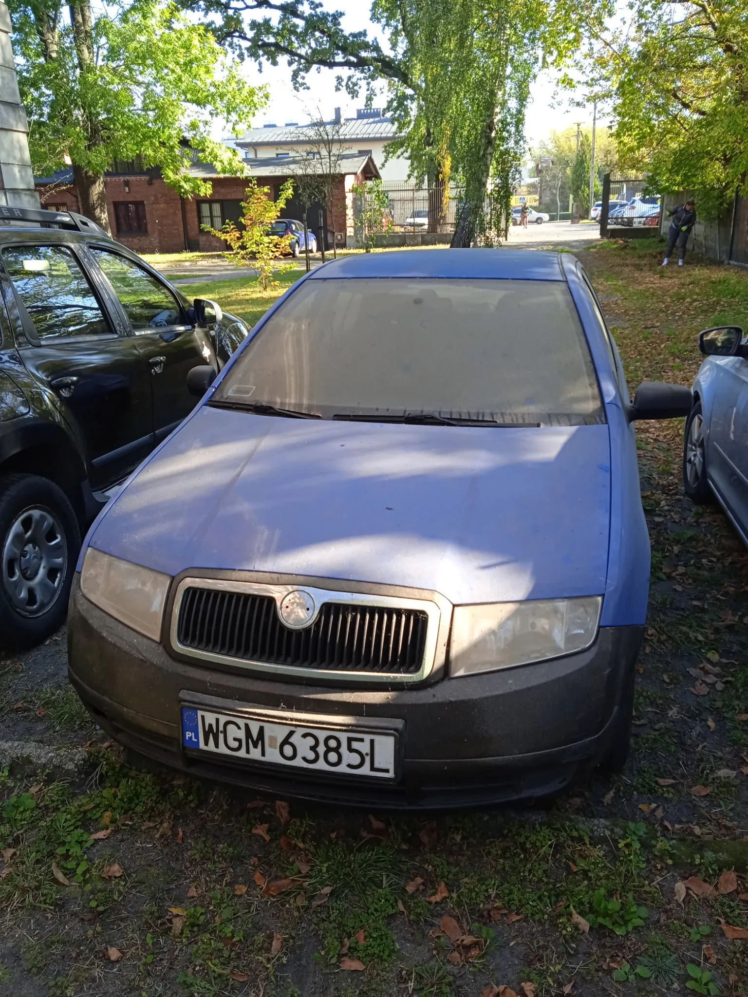 Niebieski samochód osobowy marki Skoda zaparkowany na trawiastym terenie. Samochód ma zmatowioną przednią szybę oraz czarny przedni zderzak. Tablica rejestracyjna samochodu to WGM 6385L. Po lewej stronie widoczny jest fragment ciemnego samochodu terenowego, a po prawej fragment srebrnego auta. W tle znajdują się drzewa oraz budynek mieszkalny.