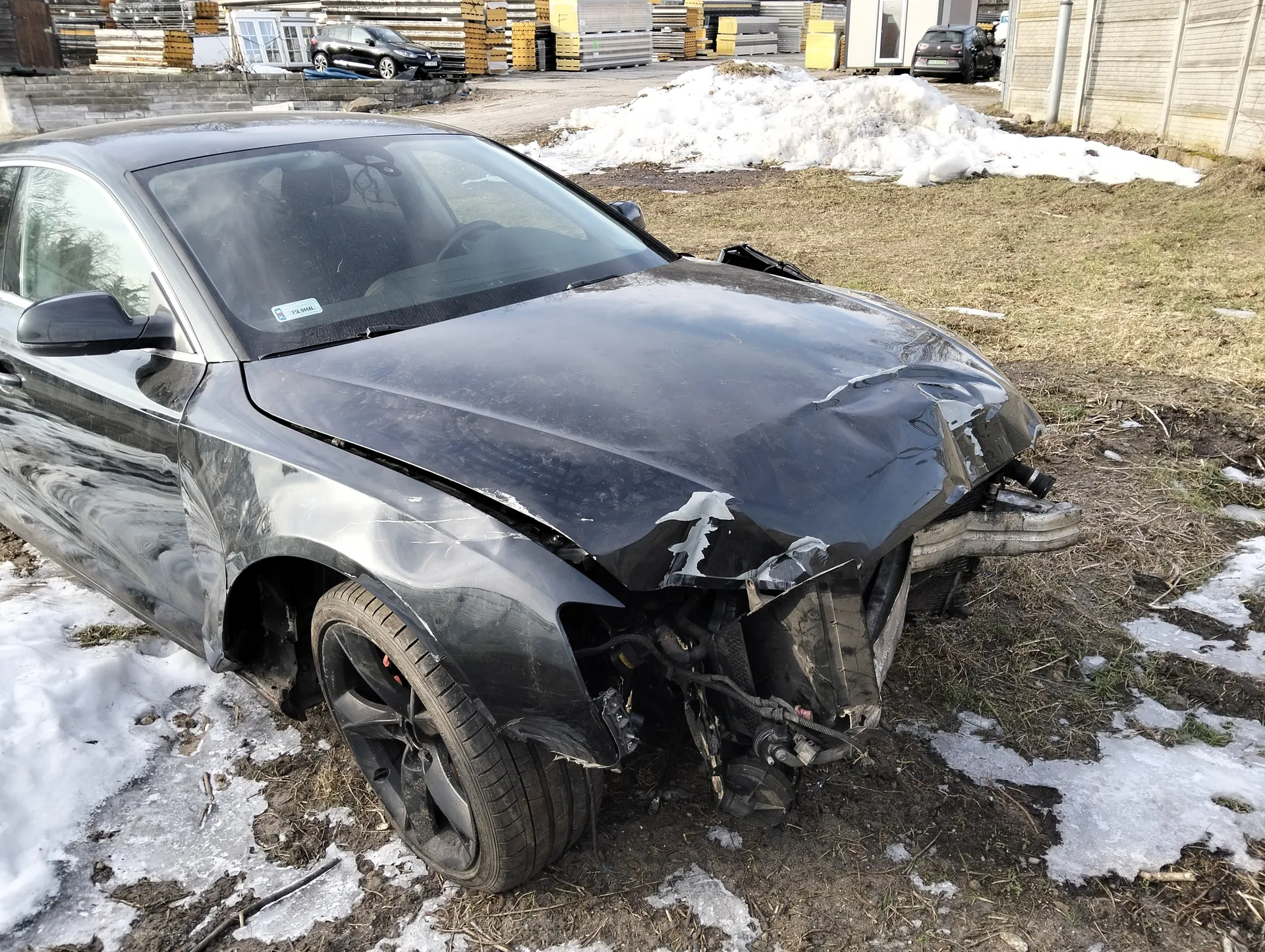 Zniszczony czarny samochód osobowy z uszkodzonym przodem, w tym zdeformowaną maską, zniszczonym zderzakiem i odsłoniętymi częściami silnika. Samochód stoi na terenie z resztkami śniegu i suchą trawą. W tle widoczne są inne pojazdy, stosy drewnianych palet, budynki oraz duża kopa śniegu. Na przedniej szybie samochodu znajduje się mały nieczytelny naklejony znaczek.