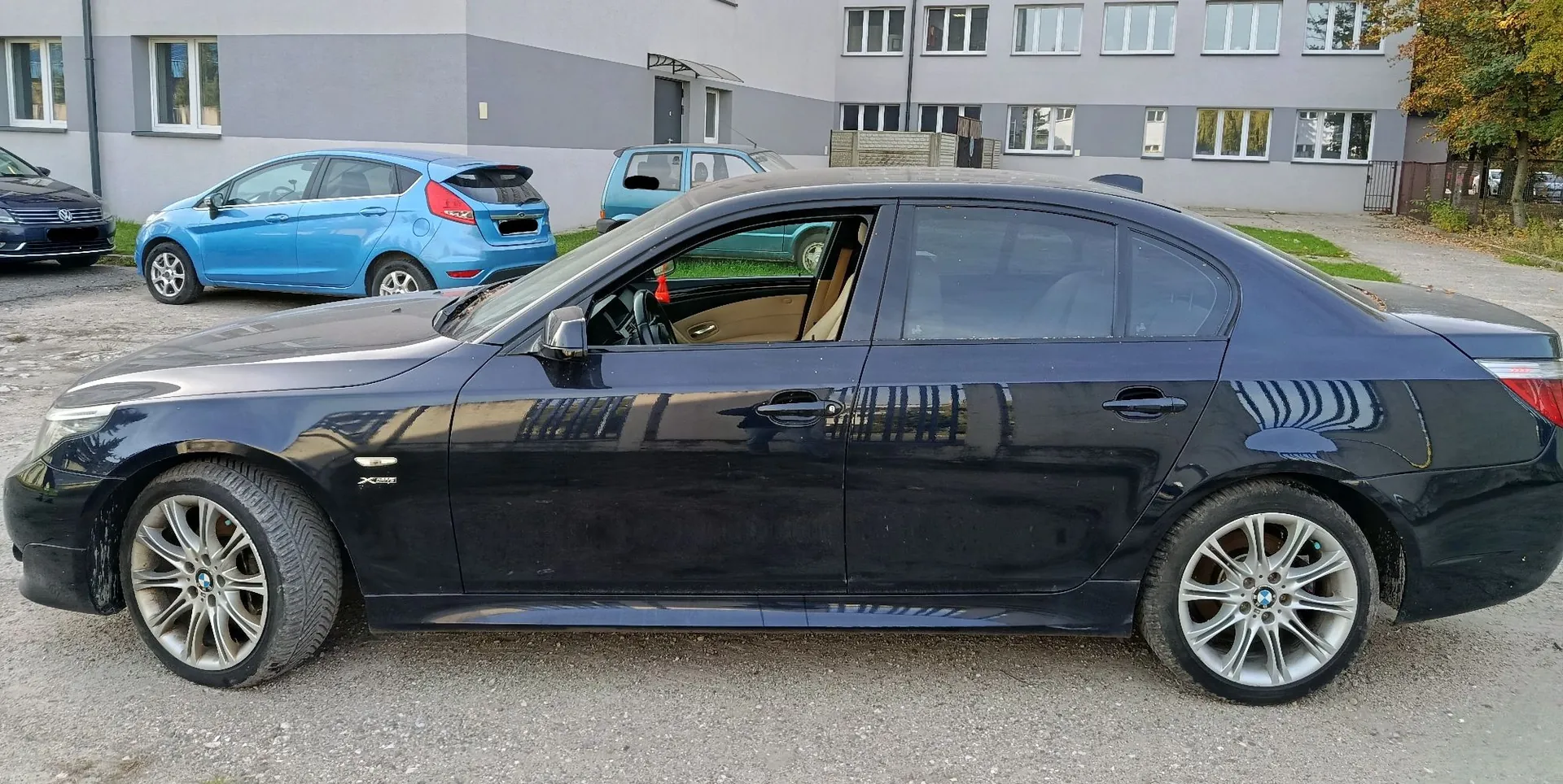 Czarny samochód osobowy typu sedan zaparkowany bokiem na żwirowym podłożu. W tle widoczny jest niebieski samochód kompaktowy oraz szary, wielopiętrowy budynek z wieloma oknami.