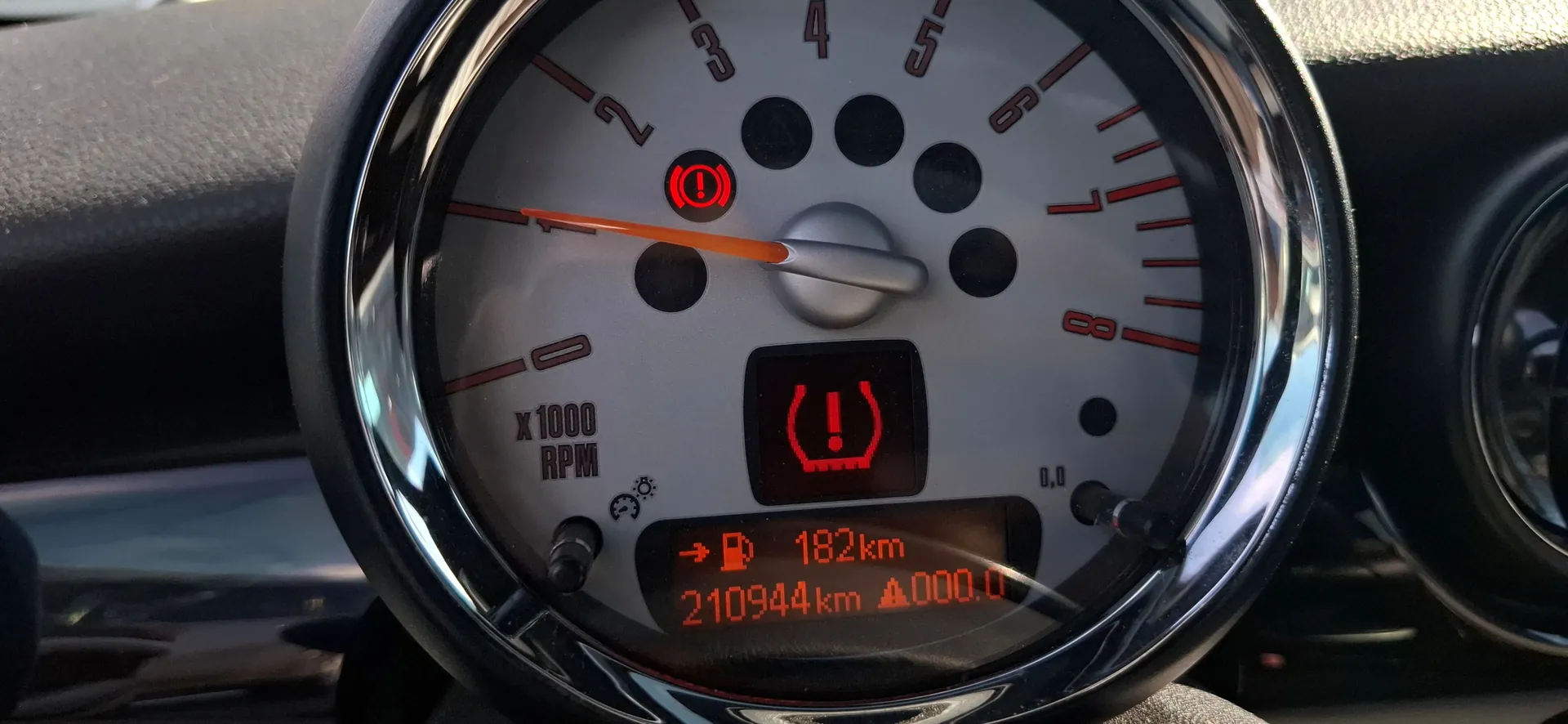 Zbliżenie na wskaźnik obrotomierza w samochodzie. Na tarczy widoczna jest skala od 0 do 8 x 1000 RPM. Pomarańczowa wskazówka znajduje się w pozycji poziomej. Na tarczy świecą się dwie czerwone kontrolki ostrzegawcze: symbol wykrzyknika w kółku oraz symbol wykrzyknika wewnątrz nawiasów przypominających klocki hamulcowe. Na dole znajduje się cyfrowy wyświetlacz z tekstem: '-> ⛽ 182km' oraz '210944km ⏵ 000.0'.