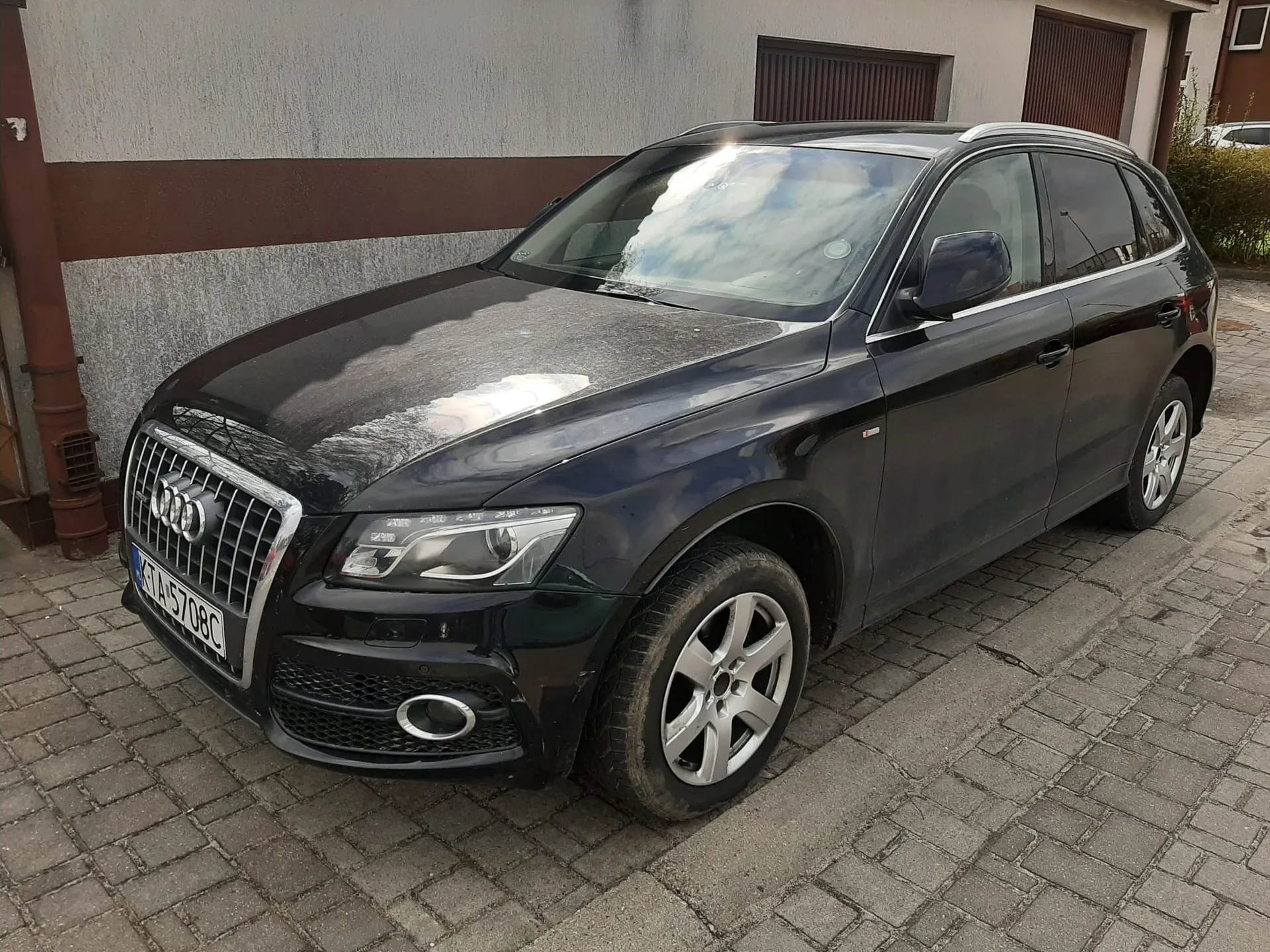 Czarny samochód Audi typu SUV zaparkowany na brukowanej powierzchni. Widoczna tablica rejestracyjna KTA 5708C. W tle budynek z szarą i brązową ścianą oraz bramami garażowymi.