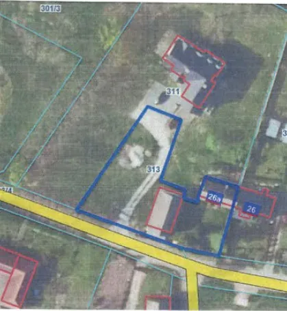 Zdjęcie lotnicze terenu z naniesionymi niebieskimi i czerwonymi liniami wyznaczającymi granice działek. Na obrazie widoczne są numery działek: 301/3 w lewym górnym rogu, 311 w górnej części, 313 w centrum oraz 26a i 26 po prawej stronie. Przez dolną część obrazu przebiega żółta droga. Teren jest zalesiony z widocznymi budynkami i podjazdami.