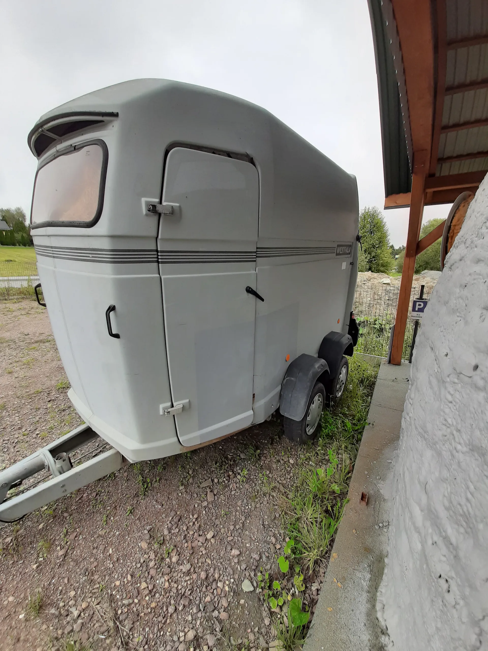 Szara przyczepa z czarnymi pionowymi liniami na boku, oknem i drzwiami z klamką. Obok przyczepy znajdują się dwa opony. W tle widoczny budynek z drewnianym daszkiem, biała ściana, trawa, żwirowa droga oraz zachmurzone niebo. Na ścianie budynku umieszczony jest niebieski znak parkingowy z literą P.