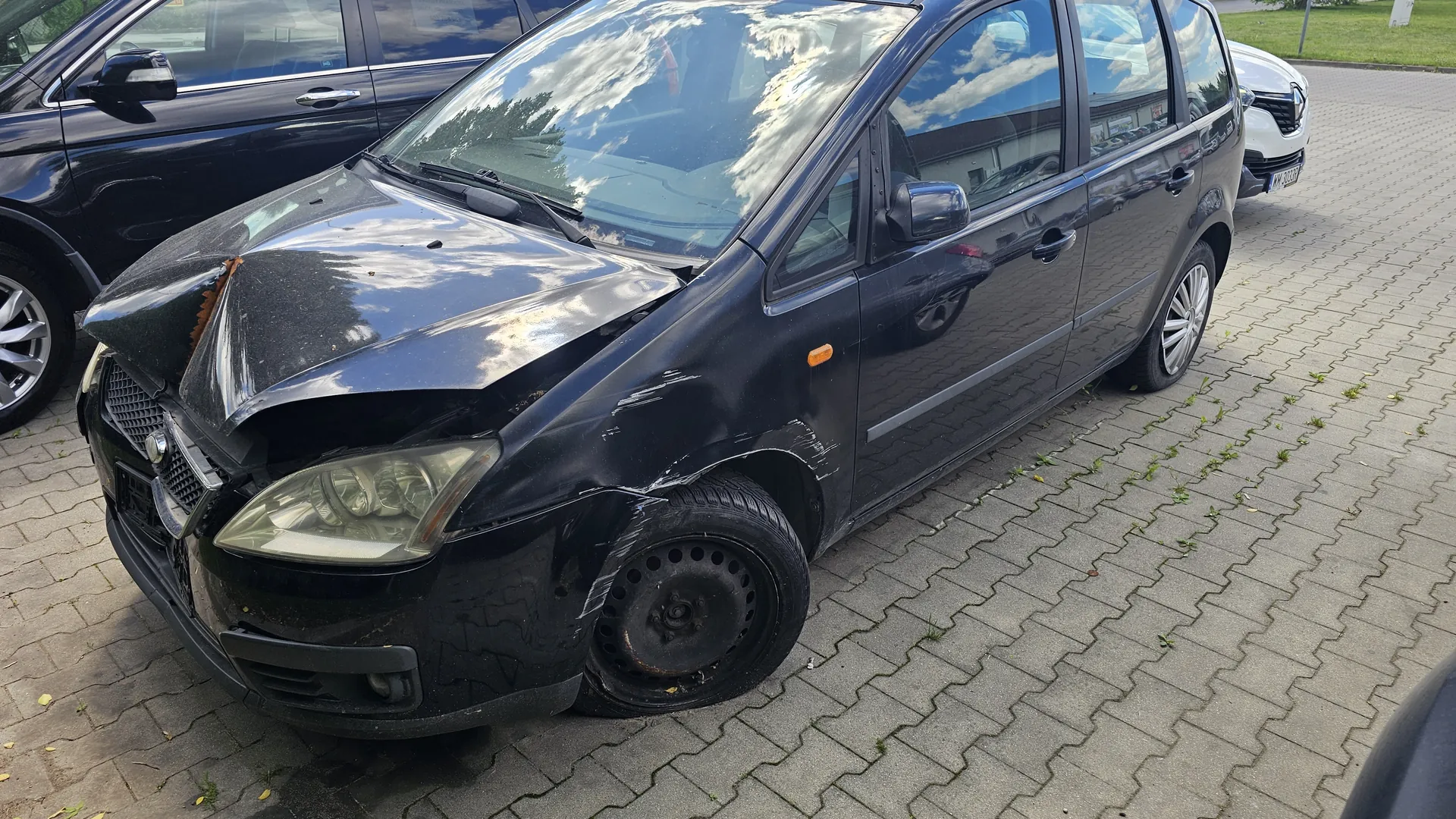 Czarny samochód osobowy z widocznymi uszkodzeniami na przodzie: zgnieciona maska, uszkodzona przednia lewa strona, brak przedniego lewego koła (zamiast niego koło zapasowe). Samochód zaparkowany na brukowanej powierzchni. W tle częściowo widoczne inne pojazdy: czarny samochód po lewej i biały po prawej stronie. Na niebie widoczne chmury, odbijające się w szybach pojazdu.