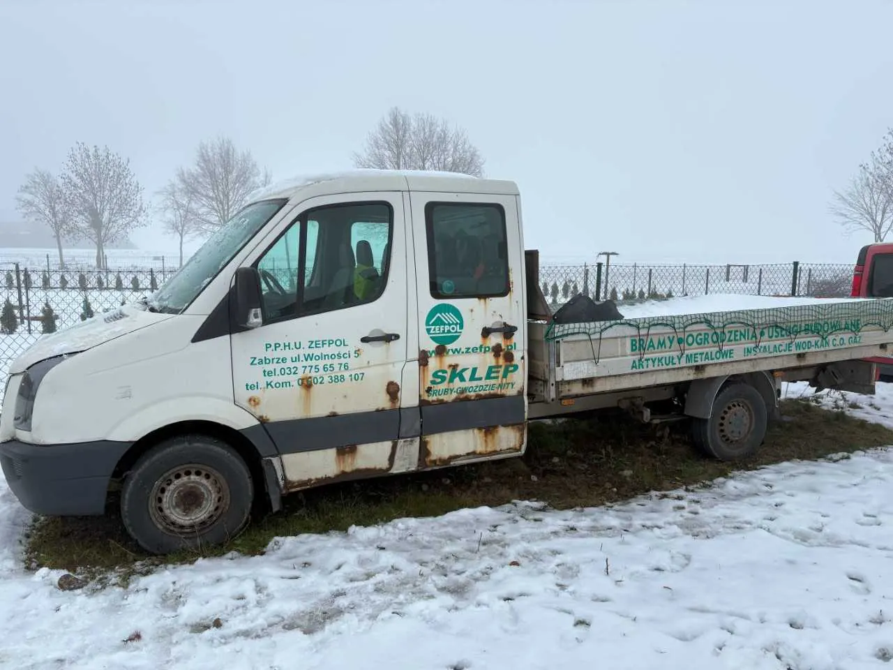 Biały samochód dostawczy z naczepą płaską, zaparkowany na śnieżnej powierzchni. Na boku pojazdu widoczny zielony tekst: 'P.P.H.U. ZEFPOL, Zabrze ul. Wolności 5, tel. 032 775 65 76, tel. Kom. 0 502 388 107', logo 'ZEFPOL' oraz 'SKLEP RURY-ODWODNIENIE'. Na naczepie napisy: 'BRAMY • OGRODZENIA • USŁUGI BUDOWLANE' i 'ARTYKUŁY METALOWE INSTALACJE WOD-KAN C.O. GAZ'. Samochód ma rdzewe plamy na boku. Tło: nagie drzewa, ogrodzenie i pochmurne niebo. Śnieg pokrywa ziemię i części pojazdu.