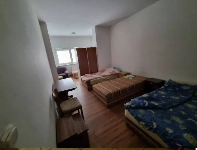 Pokój z trzema łóżkami: jednym z niebieską narzutą w wzory, drugim z paskowaną narzutą i trzecim z różowo-białą narzutą. W pokoju znajduje się stolik z dwoma krzesłami, szafa, okno oraz kanapa blisko okna. Podłoga z drewnianych desek, jasne ściany. Brak widocznego tekstu.