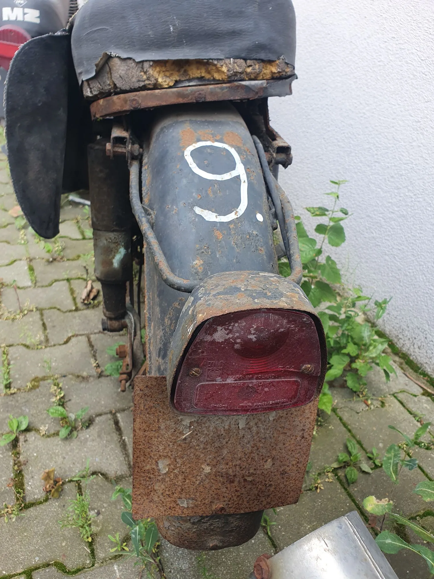 Zbliżenie na tylną część starego, zardzewiałego motocykla. Widoczny jest czarny błotnik z namalowaną białą cyfrą '6', czerwone tylne światło oraz mocno skorodowany, metalowy uchwyt tablicy rejestracyjnej. W tle widać fragment białej ściany oraz kostkę brukową z wyrastającą trawą.
