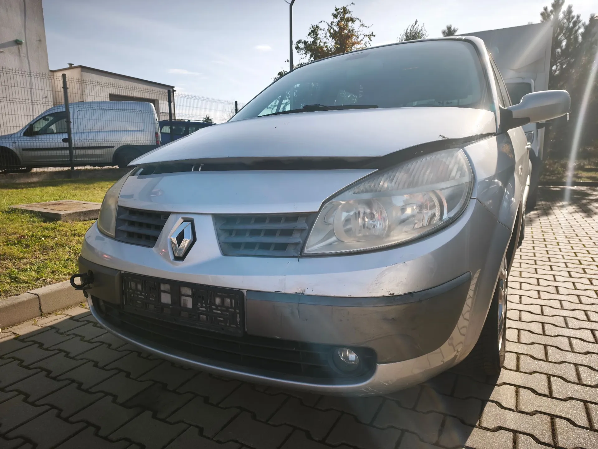 Srebrny samochód marki Renault zaparkowany na kostce brukowej. Widoczny jest przód pojazdu z logo producenta na grillu, reflektorami oraz miejscem na tablicę rejestracyjną. W tle znajduje się biały furgon, ogrodzenie z siatki oraz budynek.