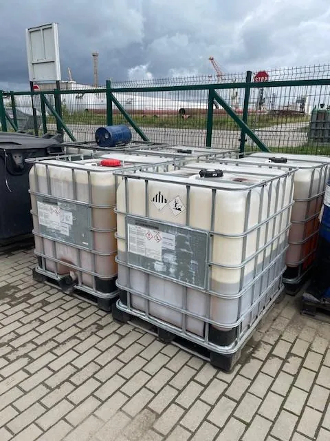 Kilka białych zbiorników IBC w metalowych klatkach, ustawionych na betonowej kostce brukowej. Zbiorniki zawierają jasną ciecz i posiadają etykiety ostrzegawcze. W tle widoczny jest zielony płot z siatki, za którym znajduje się biały samolot oraz żurawie portowe pod zachmurzonym niebem.