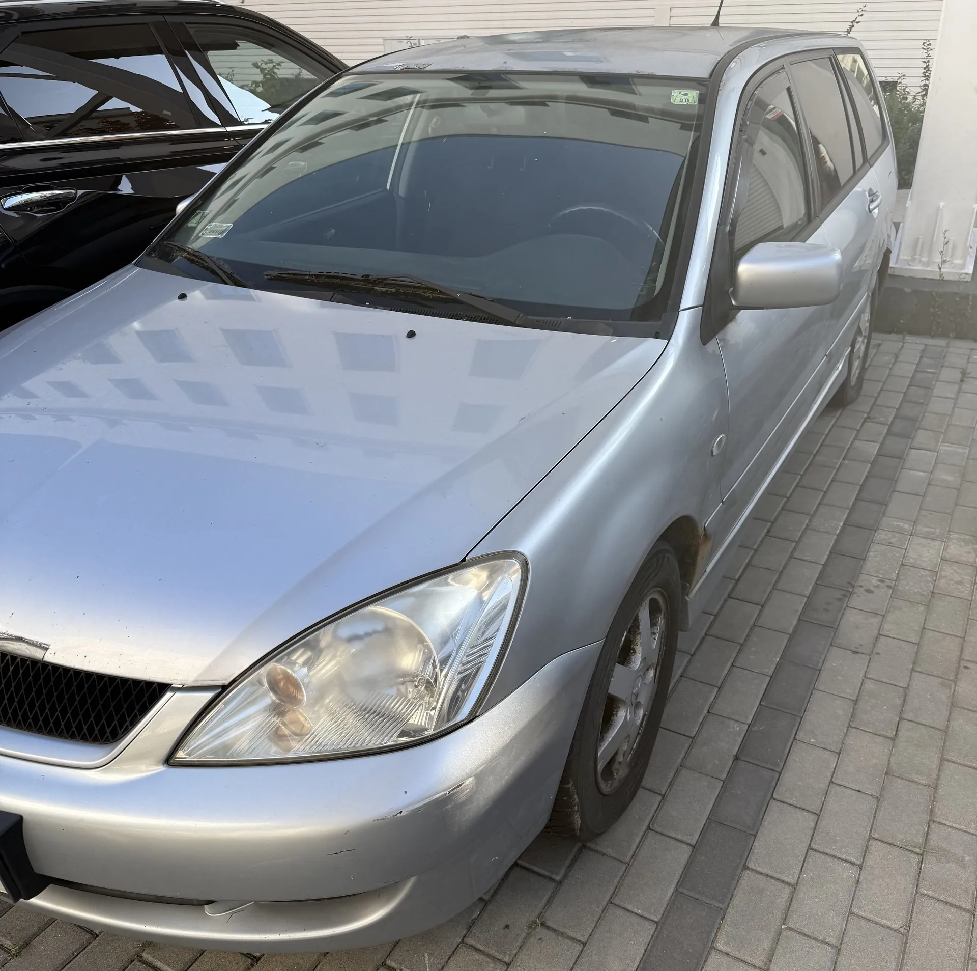 Srebrny samochód typu kombi zaparkowany na kostce brukowej. Widoczny jest przód i prawy bok pojazdu, w tym reflektor, maska, przednia szyba oraz lusterko boczne. Obok stoi inny, czarny samochód.