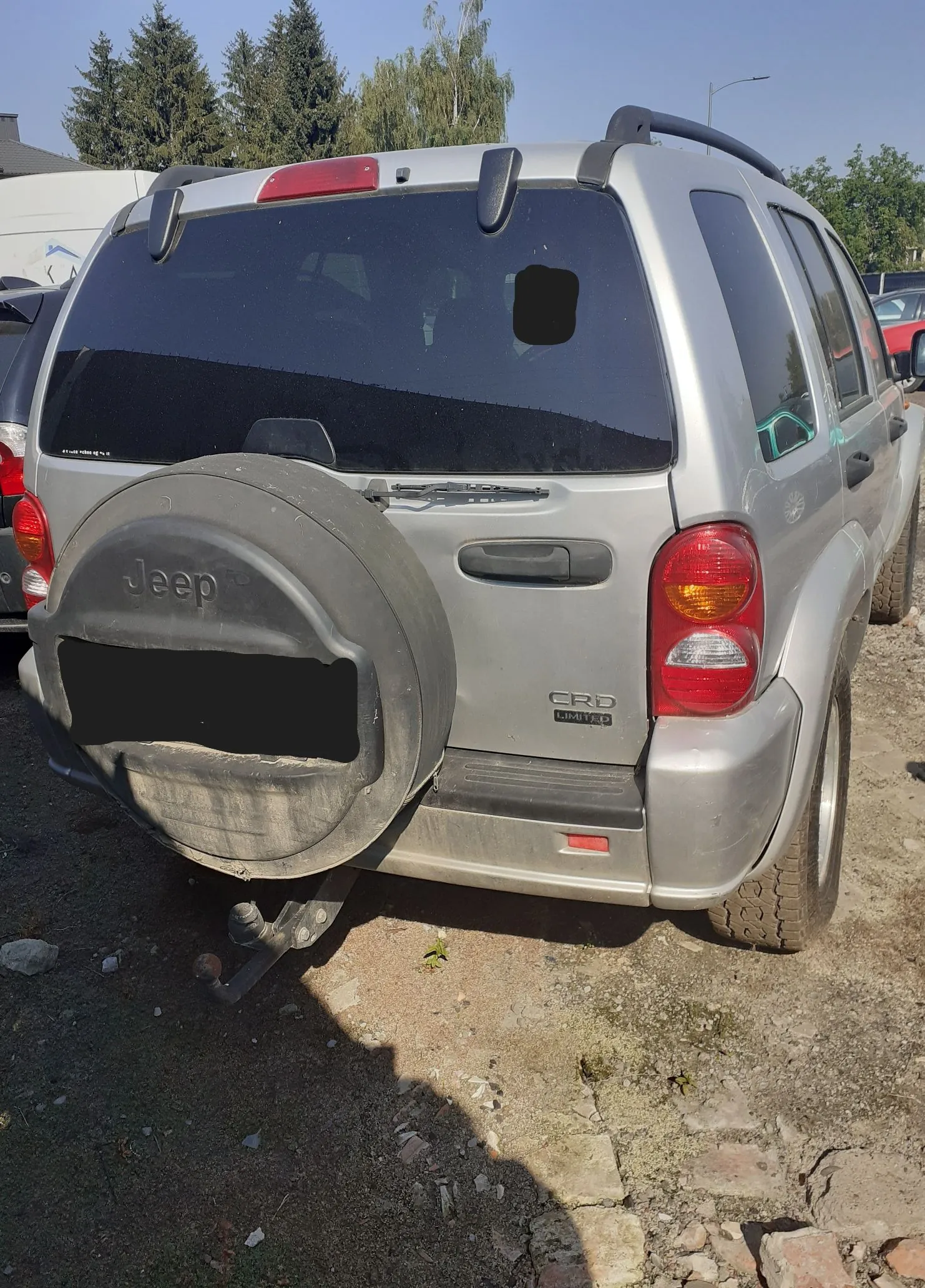 Srebrny samochód marki Jeep z tyłu, zaparkowany na piaszczystym podłożu. Pojazd posiada zapasowe koło zamontowane na klapie bagażnika z napisem 'Jeep', a poniżej znajduje się zakryta tablica rejestracyjna. Po prawej stronie widnieje napis 'CRD LIMITED'. Samochód ma ciemne szyby i zamontowany hak holowniczy.