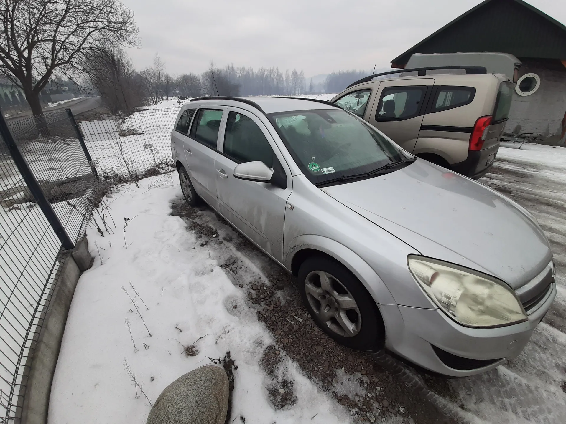 Dwa samochody zaparkowane na śnieżnej powierzchni. Przedni srebrny samochód typu kombi z zielonym naklejką na przedniej szybie, za nim beżowy samochód dostawczy. Po lewej stronie metalowe ogrodzenie, na ziemi śnieg i żwir. W tle drzewa bez liści, budynek z zielonym dachem oraz chmurne niebo. Na pierwszym planie duży kamień.