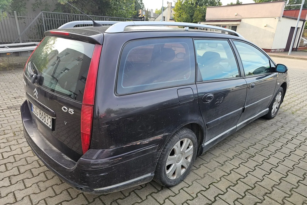 Czarny samochód marki Citroën C5 w wersji kombi, zaparkowany na kostce brukowej. Widoczny jest tył oraz prawy bok pojazdu. Samochód ma tablicę rejestracyjną o numerze ZG 86833. W tle znajduje się ogrodzenie z siatki, zielone drzewa oraz jasny budynek.
