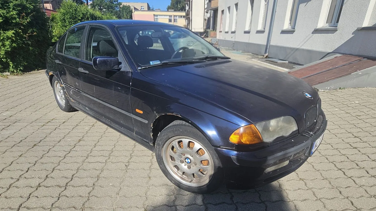 Ciemnoniebieski samochód osobowy marki BMW serii 3 (model E46) zaparkowany na kostce brukowej przed jasnym budynkiem. Auto jest widoczne z przodu i z boku, posiada widoczną rdzę na nadkolu przednim oraz zmatowiałe reflektory. W tle widać zielone krzewy i fragmenty zabudowy mieszkalnej.