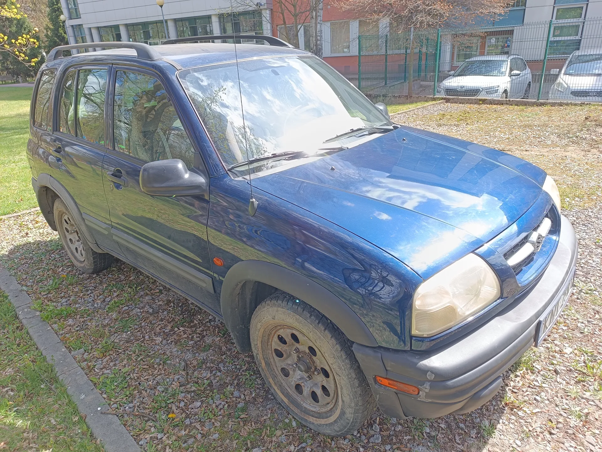 Niebieski samochód typu SUV z dachowym bagażnikiem zaparkowany na żwirowym obszarze. Po lewej stronie widoczna trawa, w tle budynek z oknami, zielone ogrodzenie oraz dwa białe samochody. Na przedniej szybie odbicie nieba i drzew. Koła mają brudne felgi. W tle drzewa bez liści i częściowo zielone krzewy.