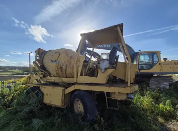 Stary, zardzewiały, żółty samochód betoniarka zaparkowany w wysokiej trawie. W tle widoczna jest żółta koparka marki Volvo. Niebo jest jasnoniebieskie z delikatnymi chmurami, a słońce znajduje się wysoko nad pojazdami.