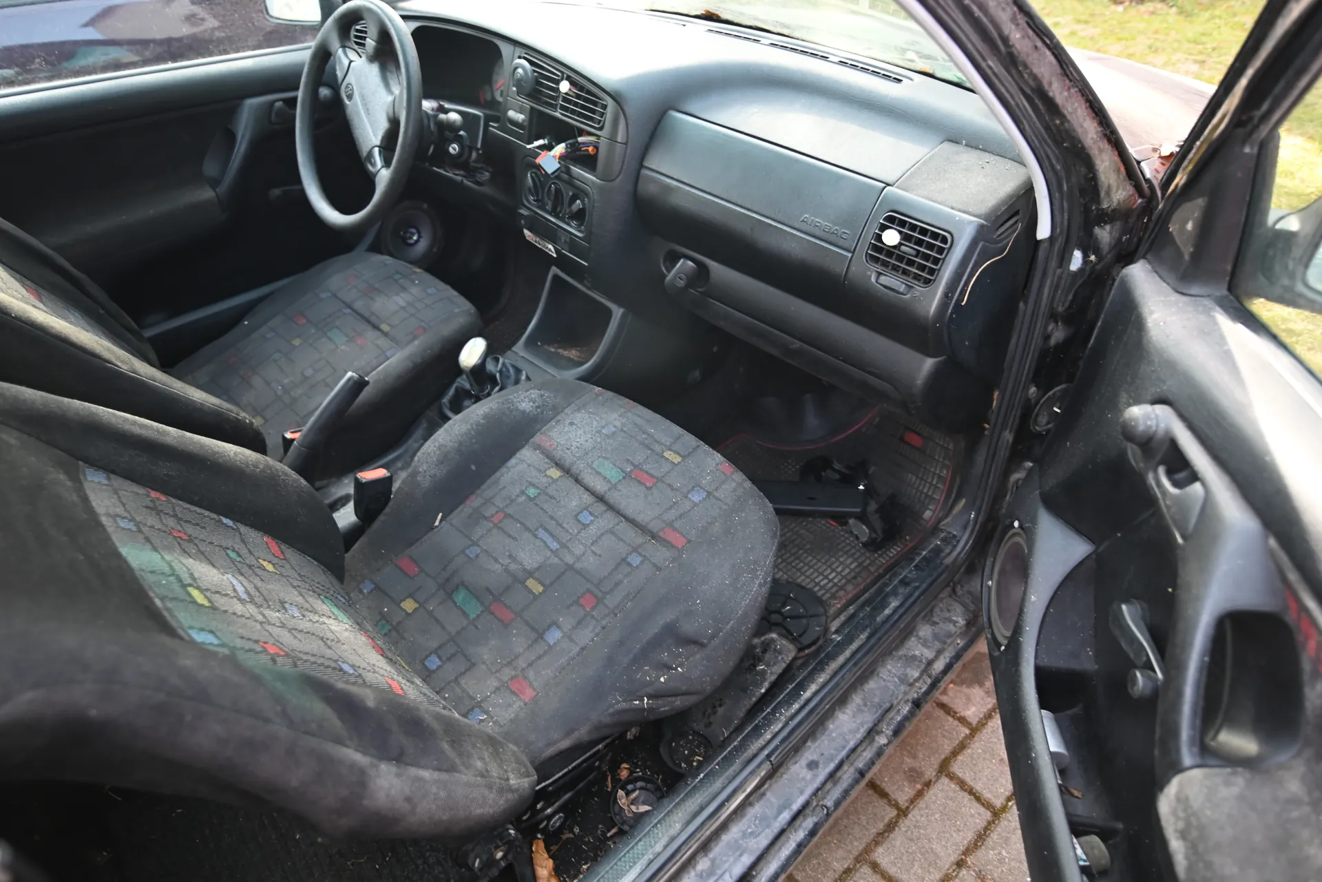Wnętrze samochodu z otwartą drzwiami kierowcy. Widoczne czarne siedzenia z kolorowym wzorem kwadratów (czerwonych, zielonych, niebieskich, żółtych), kierownica po lewej stronie, deska rozdzielcza z napisem "AIRBAG", przyciski sterujące, dźwignia zmiany biegów oraz brudne dywaniki z widocznymi śmieciem i zanieczyszczeniami na podłodze. Zewnętrzny widok przez otwarte drzwi ukazuje brukowaną nawierzchnię i trawę.