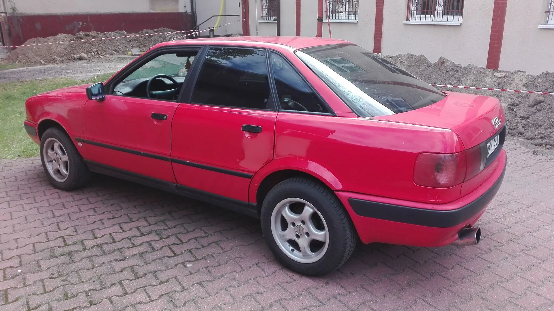 Czerwony samochód osobowy marki Audi A6 zaparkowany na kostce brukowej. Pojazd jest pokazany z profilu od strony pasażera i tyłu. W tle widoczny jest fragment białego budynku z czerwonymi detalami oraz hałda ziemi oddzielona biało-czerwoną taśmą ostrzegawczą.
