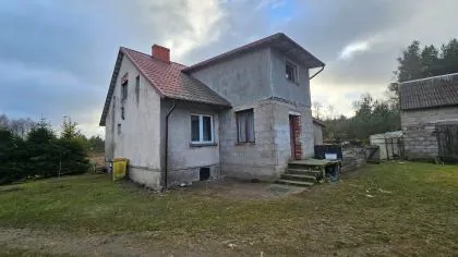 Zdjęcie jednokondygnacyjnego domu z poddaszem w stanie surowym, z szarymi ścianami z bloczków betonowych i czerwonym dachem. Budynek posiada kilka okien oraz wejście z betonowymi schodami. W tle widoczne są drzewa i zachmurzone niebo, a po prawej stronie fragment innego budynku gospodarczego.